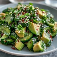 A vibrant bowl of smashed cucumber and avocado salad with sesame dressing, topped with toasted seeds and green onions.